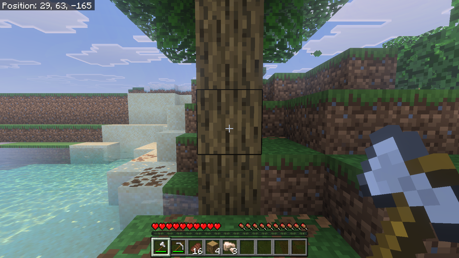 First-person Survival view aimed at a natural oak trunk beside a sandy bank; the hotbar contains four oak logs.
